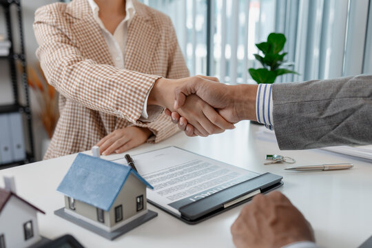 Two people in business attire finalize a real estate deal, shaking hands across a desk with house models, keys, and a contract, symbolizing agreement, trust, and successful negotiation.