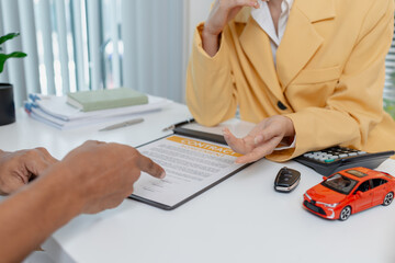 A businesswoman in a yellow suit is consulting with a male client about car purchase contracts, showing documents and car models on the desk to finalize agreement and transaction.
