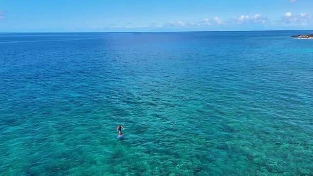 Aerial view of a lone paddle boarder gliding across the crystal-clear turquoise waters of the ocean, creating a serene contrast against the horizon, Hawi, Hawaii, United States.
