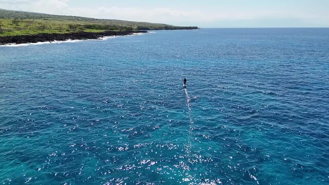 Aerial view of a lone surfer gliding across a vast expanse of blue ocean near the rocky, green coastline, creating a striking contrast, Hawi, Hawaii, United States.