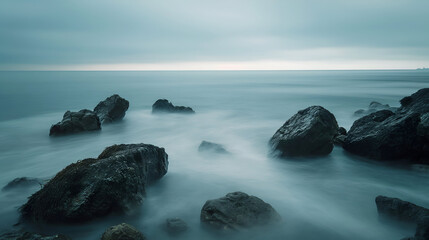 Panoramic long-exposure seascape featuring rugged coastal rocks surrounded by silky, mist-like water beneath a moody sky