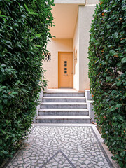 Abstract mosaic covered corridor and steps, between lush greenery, leading to pale orange door on beige walls. Travel to Athens, Greece.