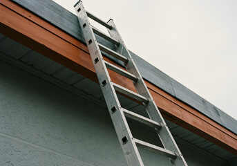 An aluminum ladder is leaning against the side of a building to reach the roof for repairs and maintenance work on the gutter