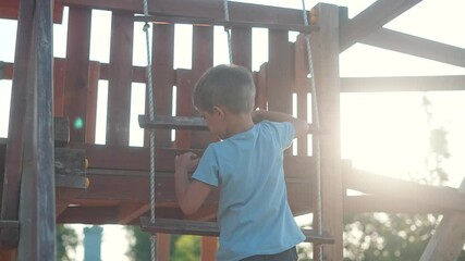 Child on playground climbs wooden rope wall. Boy uses effort and balance in summer outdoor setting. Playground climbing becomes fun activity. Climbing strengthens child motion and confidence outdoors - Powered by Adobe