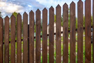 wooden fence with pointed planks stands in front of a backdrop of green trees, under a partially cloudy sky.