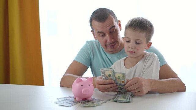 Boy looks at dollar bills while father teaches value of saving. Banking process explained slowly. Child repeats each saving step. Money used in visual finance learning guided by father support