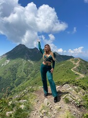 A beautiful girl in a green suit stands against the backdrop of alpine meadows with flowers in the summer in Krasnaya Polyana. The weather is clear and sunny, with a blue sky.