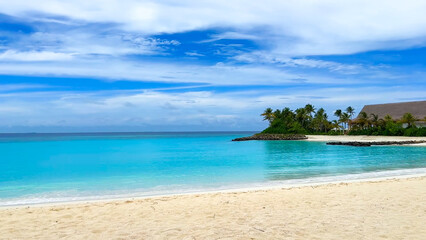 White sandy beaches meet bright turquoise waters, with a backdrop of palm trees and low resort buildings under a cloudy blue sky.