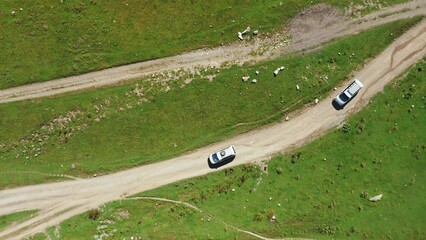 A top-down aerial view of two travelers' cars driving along the dirt mountain road one by one.