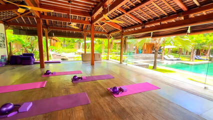 Open yoga studio with purple mats, props, wooden ceilings and glass walls overlooking the tropical pool area