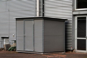 A gray storage shed stands beside a corrugated metal wall. The shed features simple design and sturdy construction. Nearby, industrial elements like vents and pipes are visible.