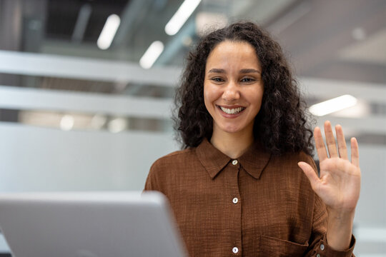 Fototapeta Young mixed-race woman smiling and waving at camera during an online video call on her laptop from a modern office, friendly and professional remote work connection