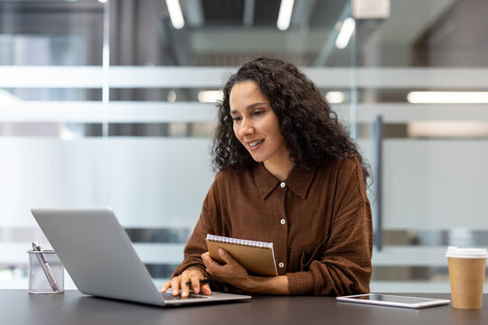 Professional young woman with curly hair working on a laptop at a modern office desk, holding a notebook and pen while smiling, expressing engagement and productivity