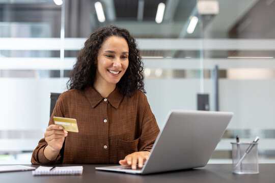 Smiling woman holding a credit card while typing on a laptop, making an online purchase or payment in a modern office setup, concentrating on the transaction