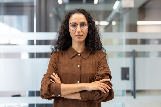 Professional businesswoman with dark curly hair and glasses standing confidently with arms crossed in a modern corporate office, representing success, management, and leadership