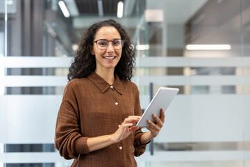 Professional businesswoman standing in a modern office, smiling confidently and holding a digital tablet, representing technology integration and success in the corporate environment