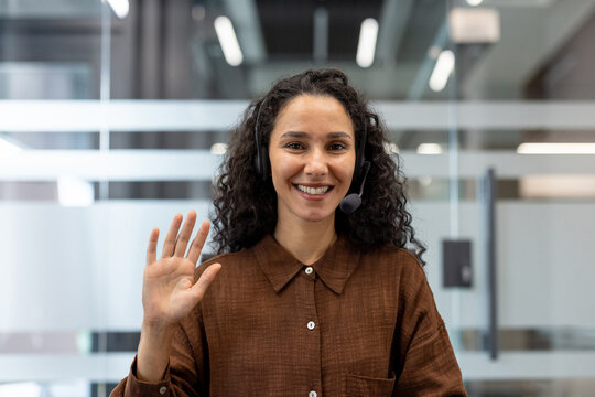 Smiling young woman wearing a headset and waving at the camera, participating in a video call or providing customer service in a modern office environment