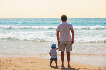 Little baby boy and his father stand on the beach and look at the sea on a sunny day in summer. The dad holds his son's hand. Rear view