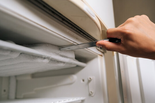 Detail cropped shot of unrecognizable man using knife to scraping away thick ice buildup from inside refrigerator, emphasizing routine task of maintaining clean kitchen appliance, close-up.