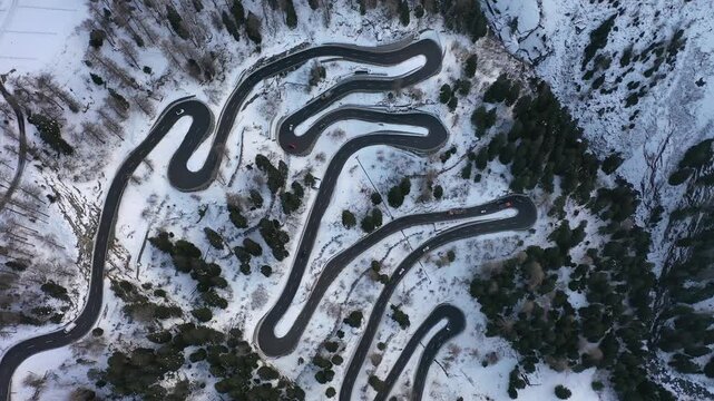 Aerial view of a winding mountain road cutting through the snowy landscape with scattered trees, creating a stark contrast, Maloja Pass, Grisons, Switzerland.