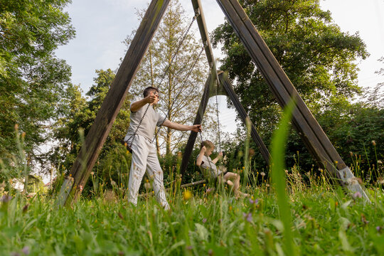 A father plays with his son on swings in a lush green park. They share laughter and joy in a beautiful natural environment. This moment reflects the bond of parenting and togetherness - Powered by Adobe