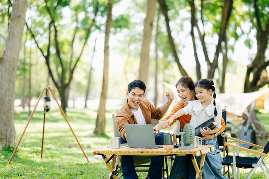 Joyful Asian family camping in a sunny garden, bonding over BBQ, tech, and laughter. Parents and daughter enjoy outdoor - Powered by Adobe