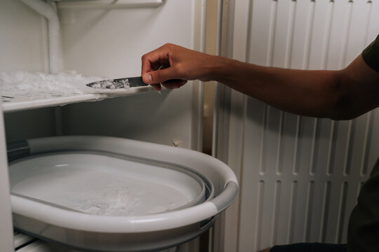 Close-up hands of unrecognizable man defrosting refrigerator, using knife to scrape off ice buildup and collecting ice chips in basin placed inside appliance for easy cleanup. Concept of domestic work