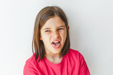 White Girl 8-9 years with long hair in a pink shirt making a funny grimace and sticking out her tongue against a white background, concept of childhood, emotions, playfulness and expression