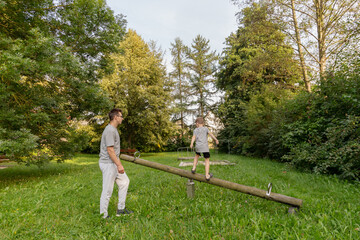 Naklejka premium A father watches as his son balances on a seesaw. They are in a vibrant park with tall trees and greenery, enjoying quality time together amidst nature