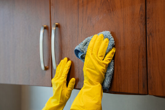 Woman in protective gloves wiping wooden cabinet surface in kitchen interior. Housekeeping and domestic chores concept. - Powered by Adobe
