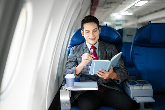 Portrait of a successful Asian businesswoman or entrepreneur in a formal suit on an airplane sitting in business class using a phone, computer laptop. Travel in style, work with grace.