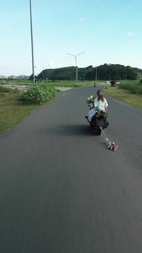 An aerial view of a newlywed couple riding a moped with a Just Married sign and tin cans tied to it. A young woman in a white dress with a bouquet sits behind the groom. A retro-style wedding.