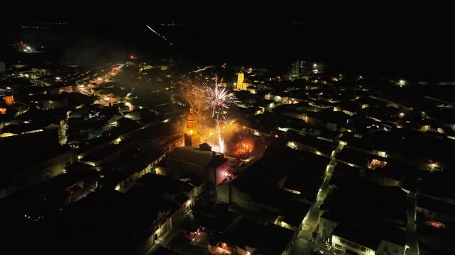 Smooth drone shot capturing fireworks lighting up the night sky over Mayorga, Spain, during El V&iacute;tor celebration, with rooftops and streets of the village illuminated below.