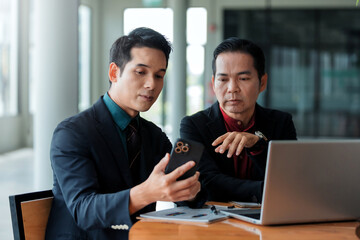 Two Asian businessmen meeting, discussing information on a smartphone, and working with a laptop in a modern office environment