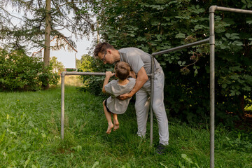 A man lifts his young son as they enjoy time together at a playground surrounded by greenery. Their laughter fills the air, highlighting the beauty of fatherhood and bonding in nature