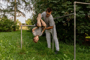 A father lifts his son upside down on playground equipment surrounded by greenery. They share...