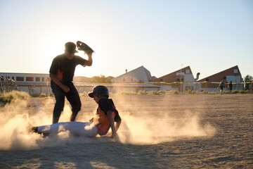 Young baseball player sliding into a dusty base as opponent waits to tag during sunset