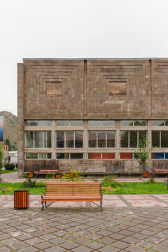 View of a blocky, textured stone building with rows of windows and a wooden bench in front, under a muted sky, Goris, Syunik Province, Armenia.