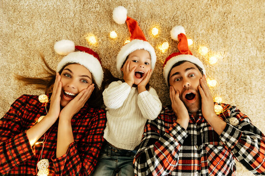 Surprised family lying on the carpet with glowing lights, parents and daughter in Santa hats holding their faces with joy, celebrating love, Christmas eve, and New Year.