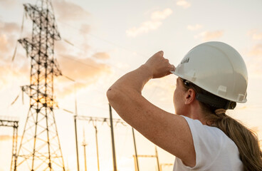 Woman in hard hat observes power lines at sunset near a utility facility while conducting site inspections