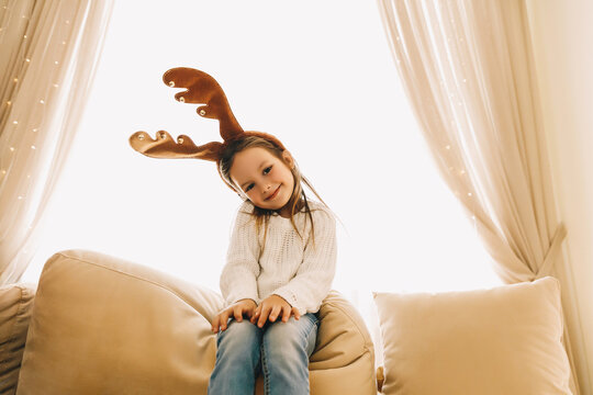 Cheerful little girl in reindeer antlers posing on sofa, dressed in cozy white sweater and jeans. Bright holiday lights and curtains behind create warm festive Christmas mood at home.