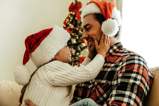 Little girl in Santa hat holding her father&rsquo;s face with love, both smiling warmly in front of a glowing Christmas tree, sharing a tender holiday moment.