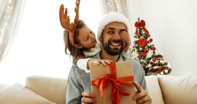 Smiling father in a Santa hat holds a Christmas gift with a red ribbon while his playful daughter in reindeer antlers covers his eye, sharing a joyful holiday moment by the tree.