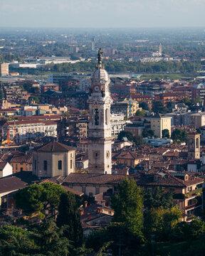 View of a tall, ornate church steeple pierces the skyline above a tapestry of terracotta roofs and verdant trees under a sunny sky, Bergamo, Lombardy, Italy.