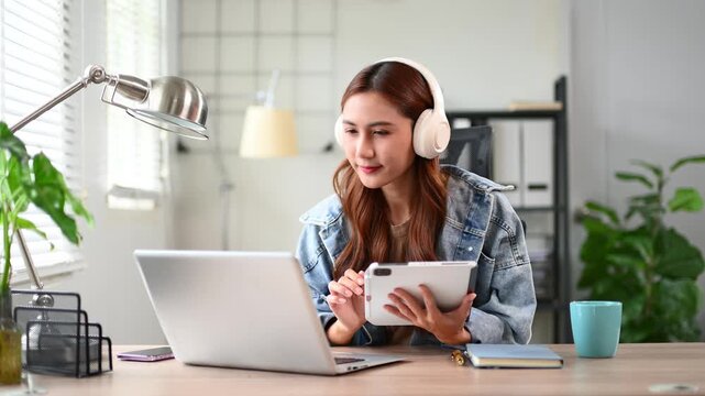 Asian woman wearing headphones working on laptop at home office desk. Remote work, online learning, and digital lifestyle concept