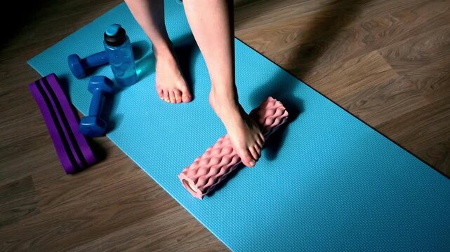 Female foot rolling massage fascia ball on yoga mat at home with dumbbells resistance band and water bottle