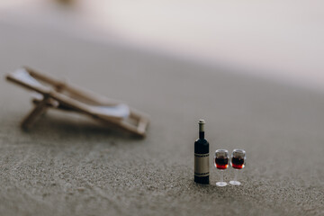 Wine glasses and a bottle of wine on the beach. A miniature wine bottle and two glasses of red wine sit on a sandy beach, with a blurred beach chair in the background.