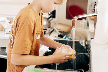 Close-up of a teenager washing a glass with detergent and foam in the kitchen sink.