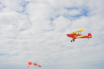 red and yellow open cockpit biplane flying in a cloudy sky above a red and white windsock, scene of an air show, concept of adventure, travel, sport © Lana Pietukhova