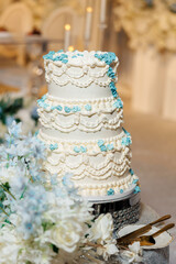 Elegant three tier white wedding cake decorated with blue flowers and ornate icing details, placed on a sparkling stand with floral arrangement and golden cutlery nearby
